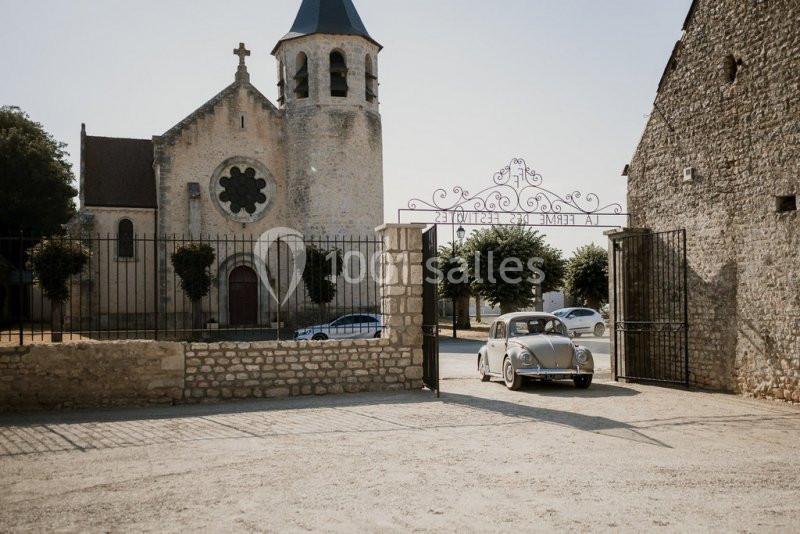 Une voiture ancienne passe devant une église en pierre et un portail ouvert dans un village ensoleillé.