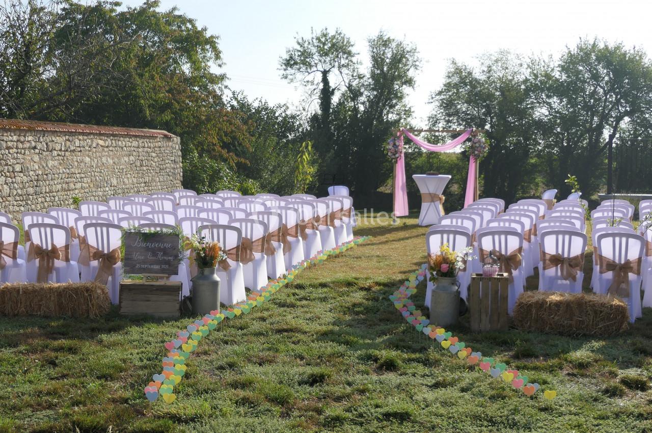 Chaises blanches alignées avec décorations champêtres, prêtes pour une cérémonie en extérieur devant une arche ornée de…