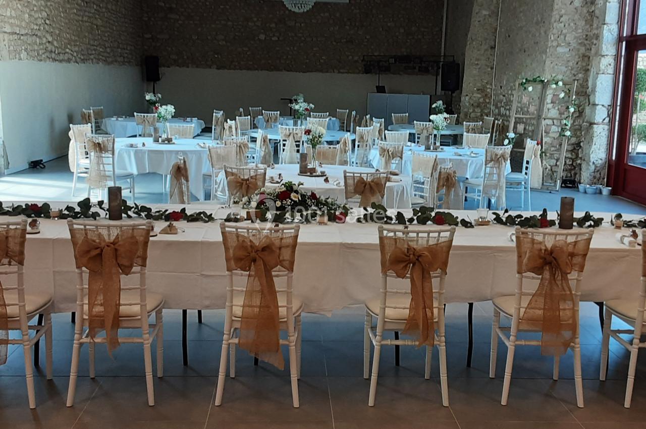 Salle de réception décorée pour un événement, avec tables dressées, chaises ornées de nœuds en toile de jute et compositions…
