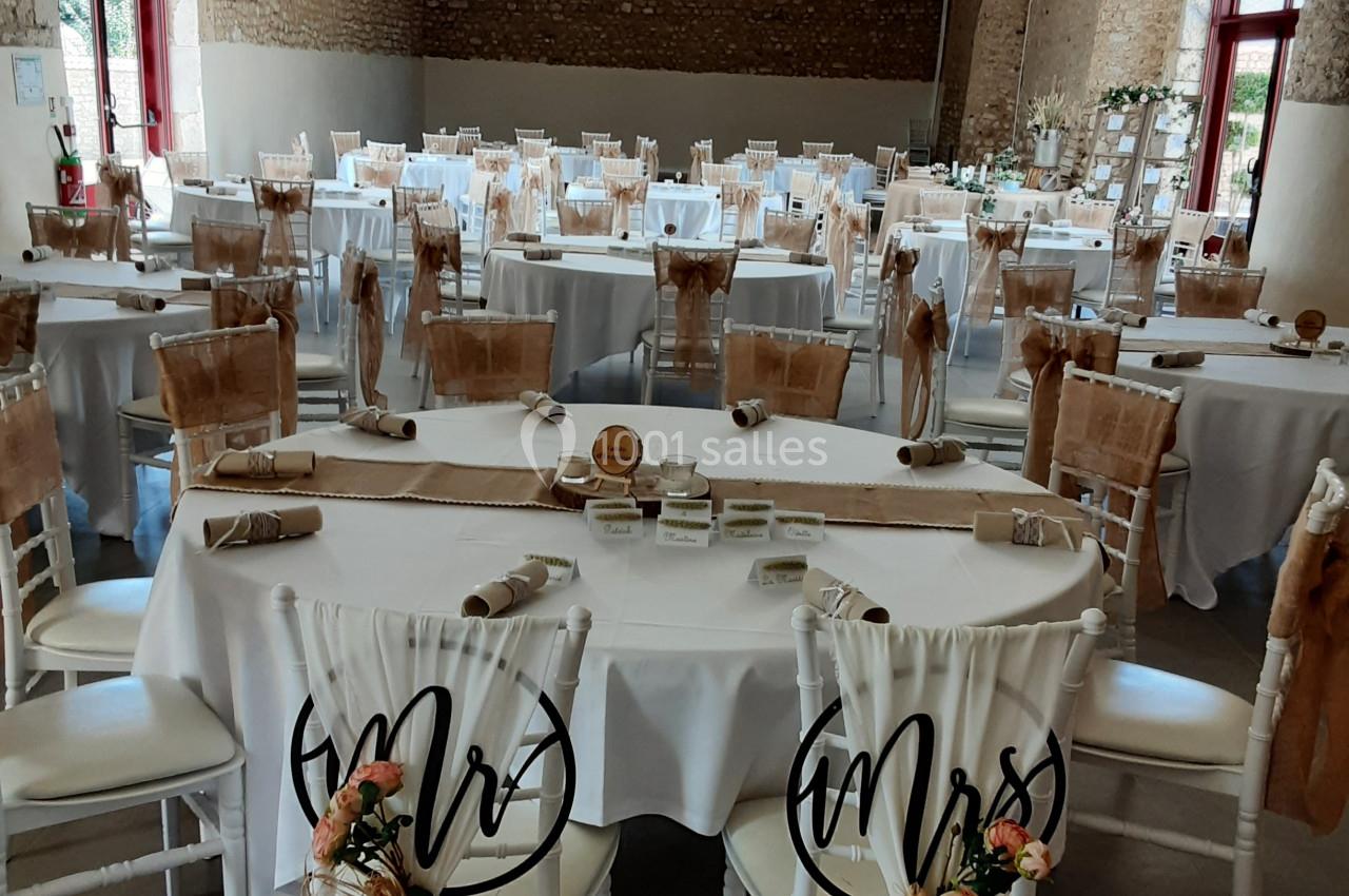 Salle de réception décorée pour un mariage, avec tables dressées, chaises ornées et lumière naturelle.
