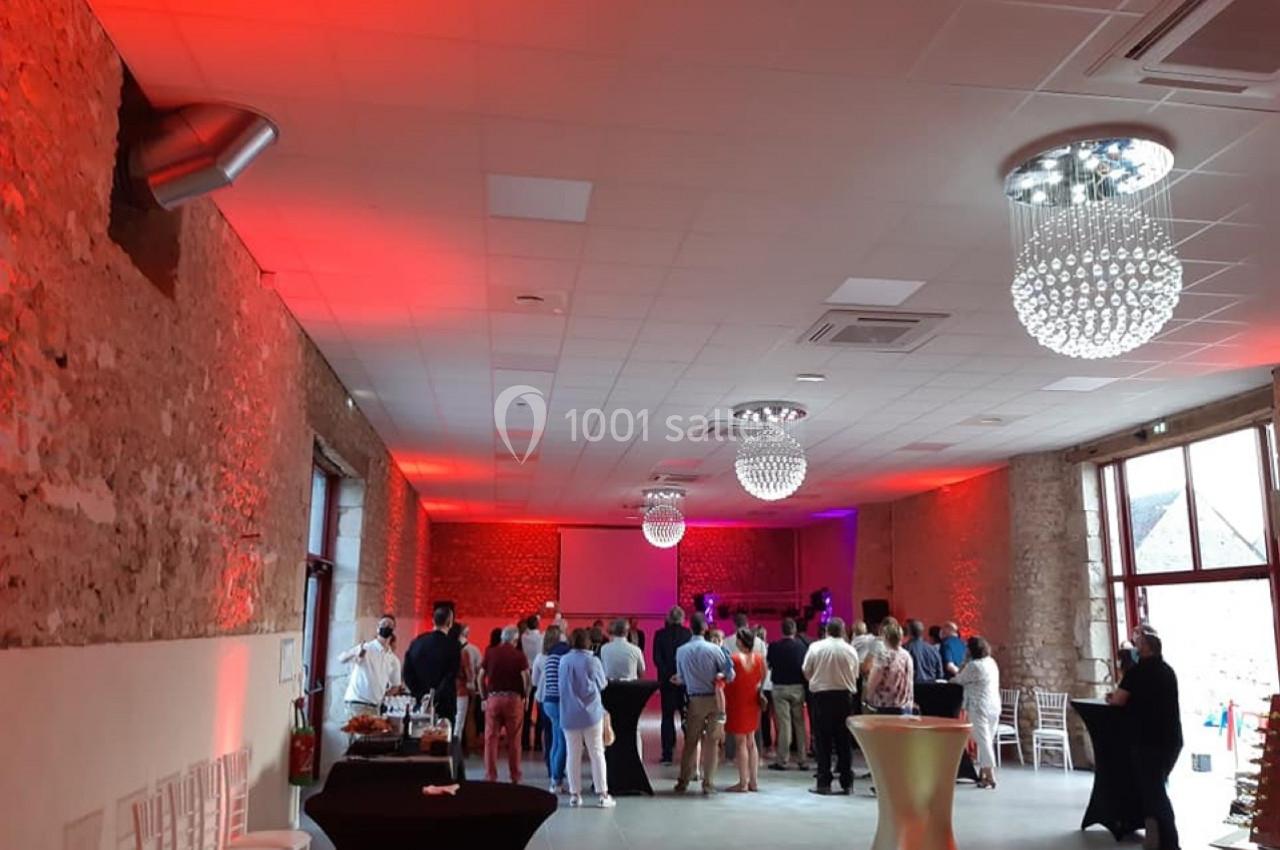 Salle de réception avec des invités debout, éclairée par des lumières rouges et violettes, et des tables hautes disposées.