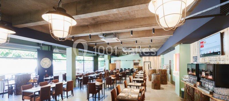 Salle de restaurant lumineuse avec tables alignées, chaises en bois, grandes fenêtres et plafond en béton apparent.