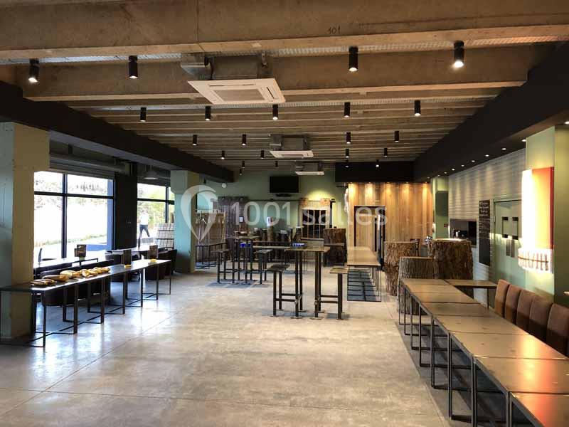 Salle spacieuse avec tables et chaises alignées, éclairage suspendu et grandes baies vitrées donnant sur l'extérieur.