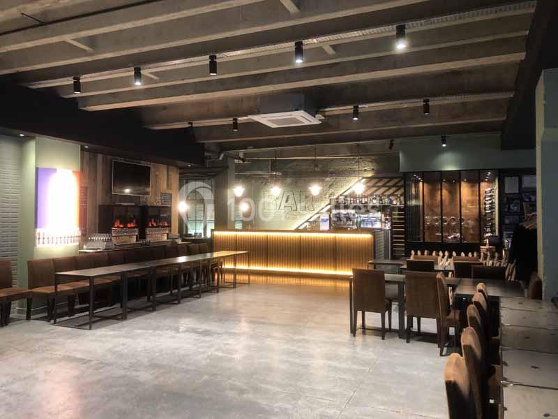 Intérieur d'un bar moderne avec un comptoir éclairé, des tables en bois et un plafond industriel.