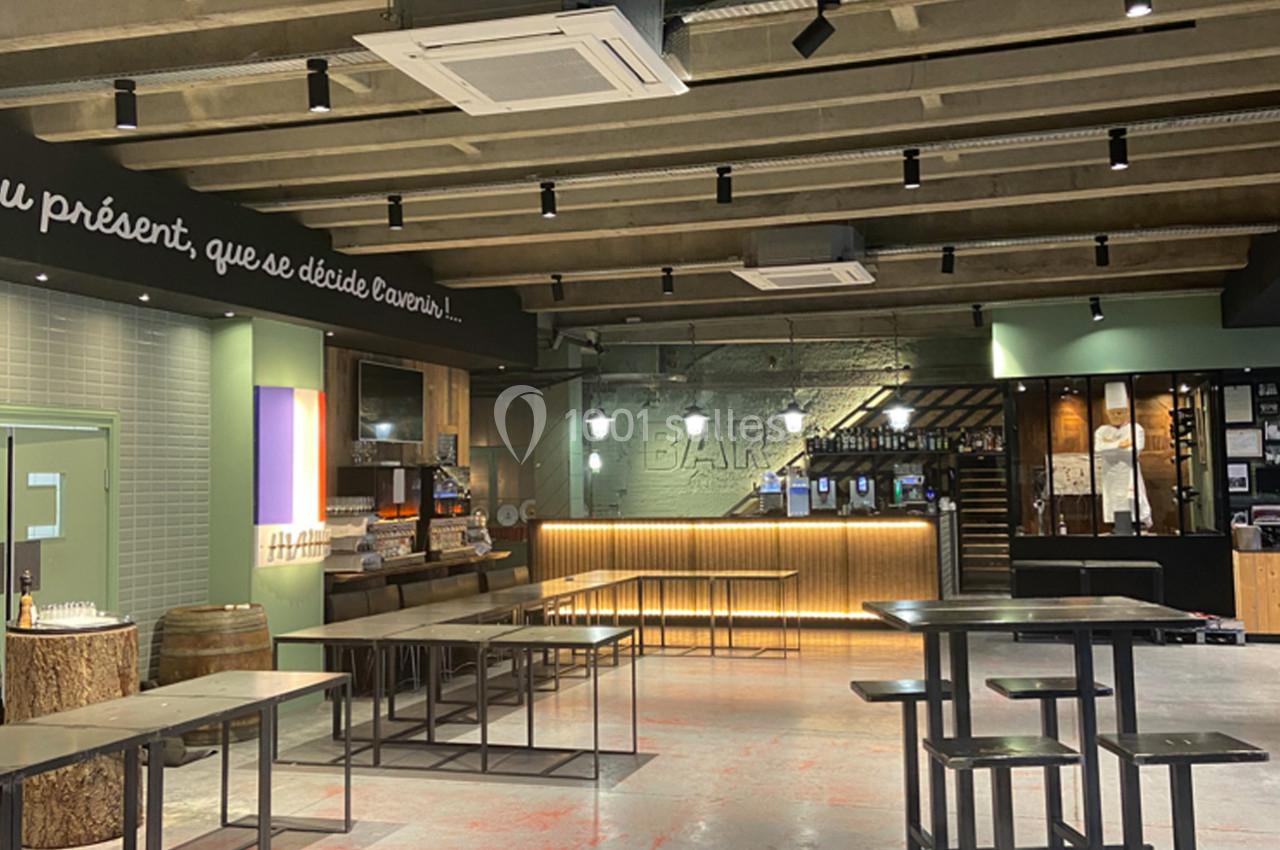 Intérieur d'un bar moderne avec tables hautes, comptoir éclairé et plafond industriel apparent.