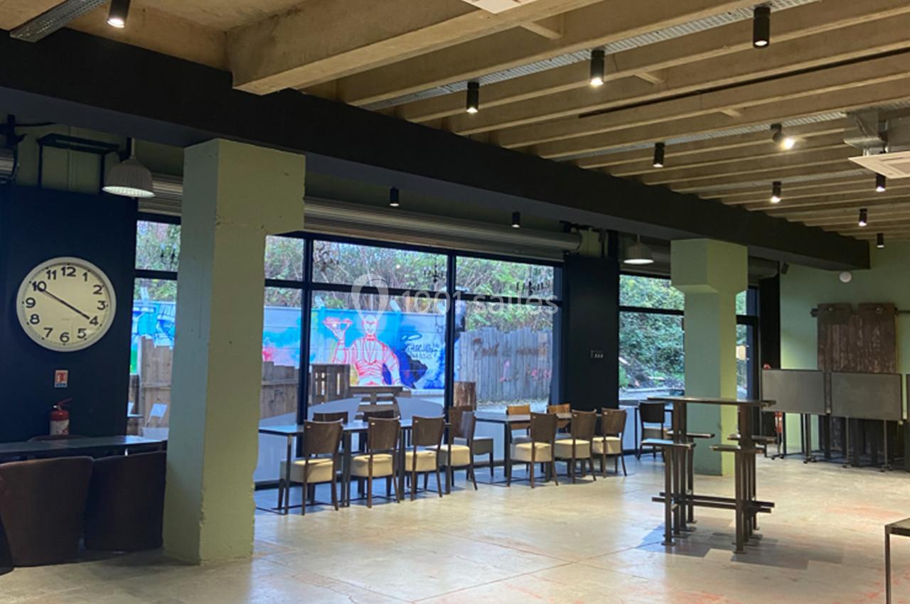 Salle lumineuse avec de grandes baies vitrées, des tables et des chaises en bois, et une horloge murale visible à gauche.