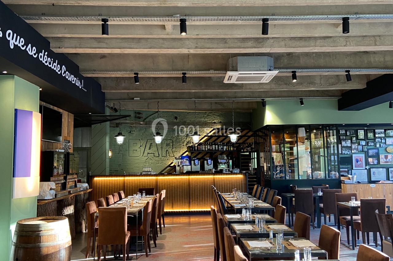 Salle de restaurant lumineuse avec tables dressées, bar en arrière-plan et plafond en béton apparent.