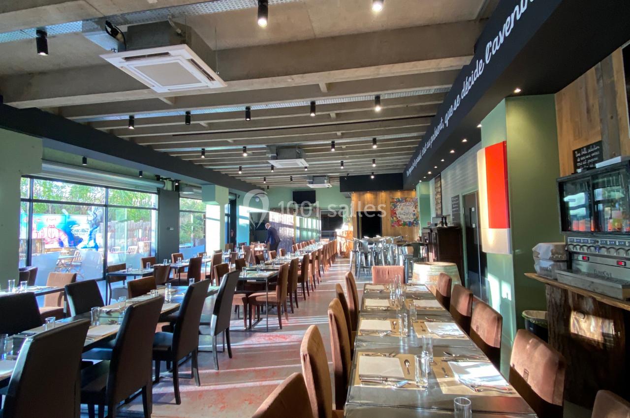 Salle de restaurant lumineuse avec tables dressées, grandes baies vitrées et plafond en béton apparent.
