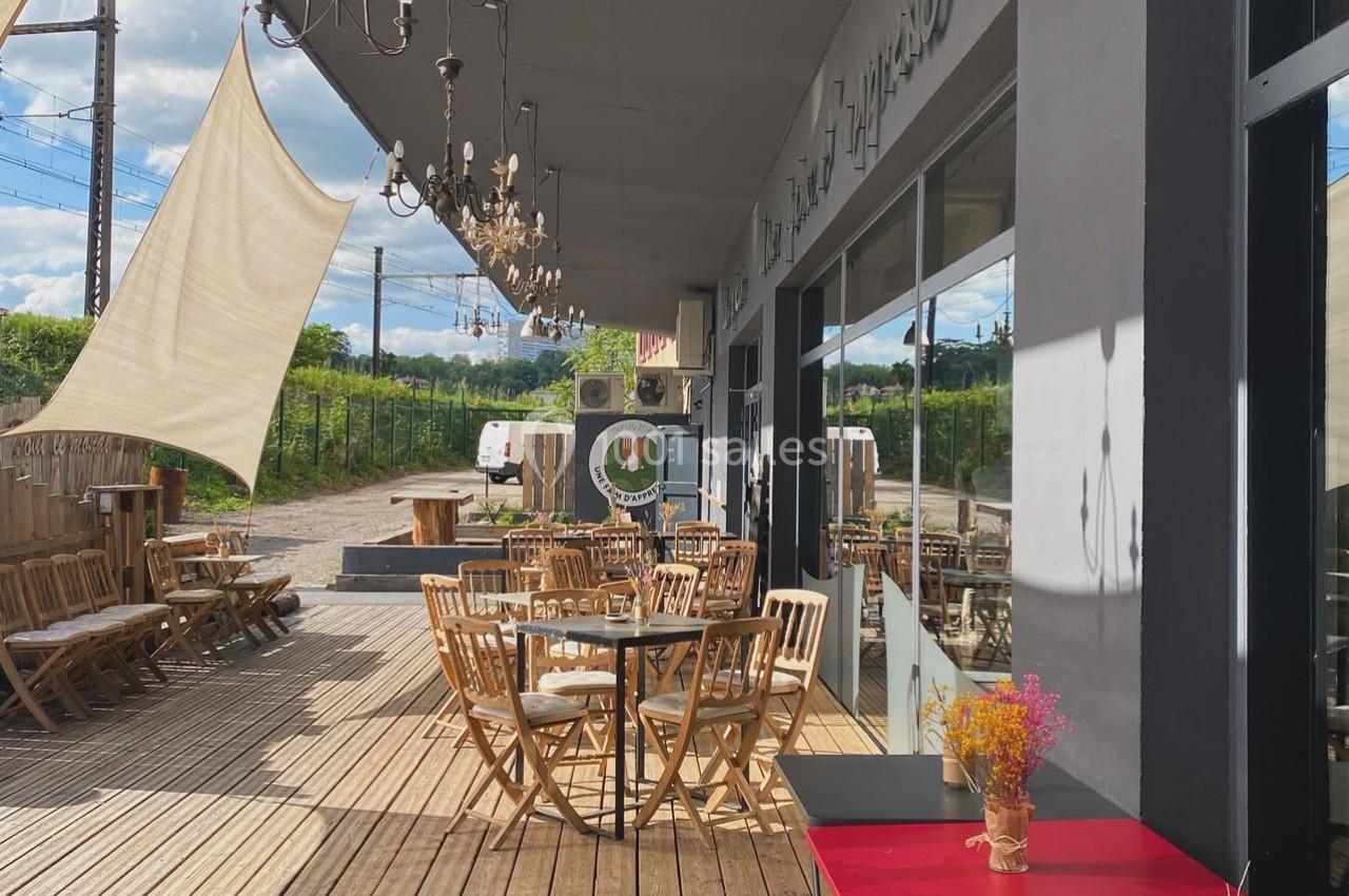 Terrasse en bois avec tables et chaises en bois, décorée de luminaires suspendus et d'une voile d'ombrage.