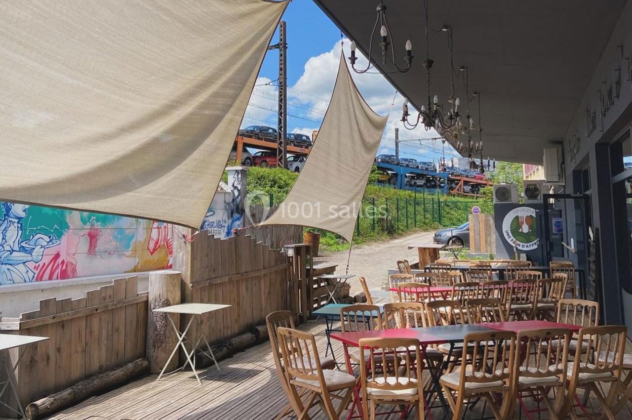 Terrasse en bois avec tables et chaises, ombragée par des voiles tendues, près d'une voie ferrée et d'un mur coloré.