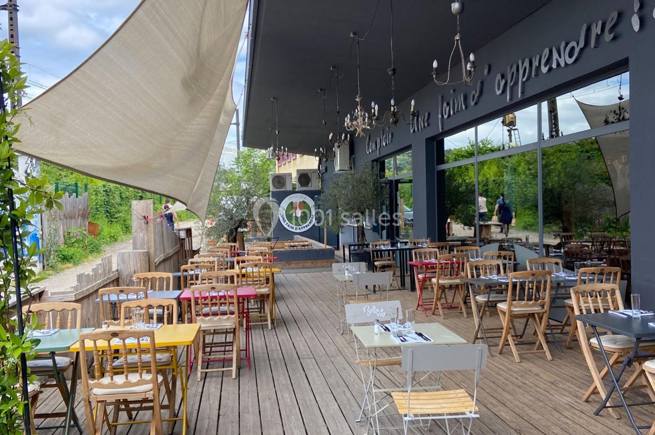 Terrasse en bois d'un restaurant avec des tables et chaises colorées, entourée de verdure et partiellement ombragée.