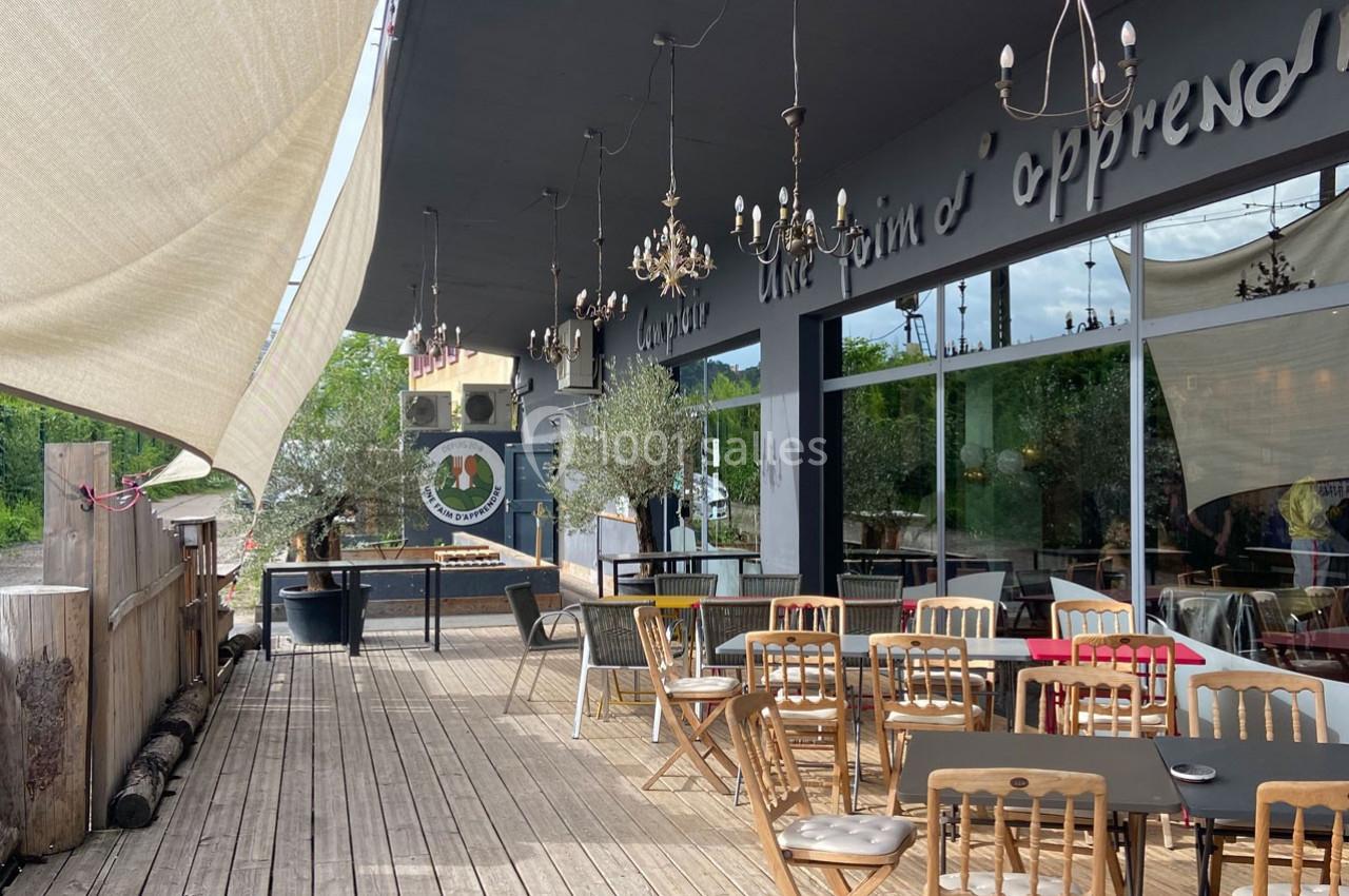 Terrasse en bois avec tables et chaises, ombragée par des voiles, devant une façade vitrée et végétation en arrière-plan.