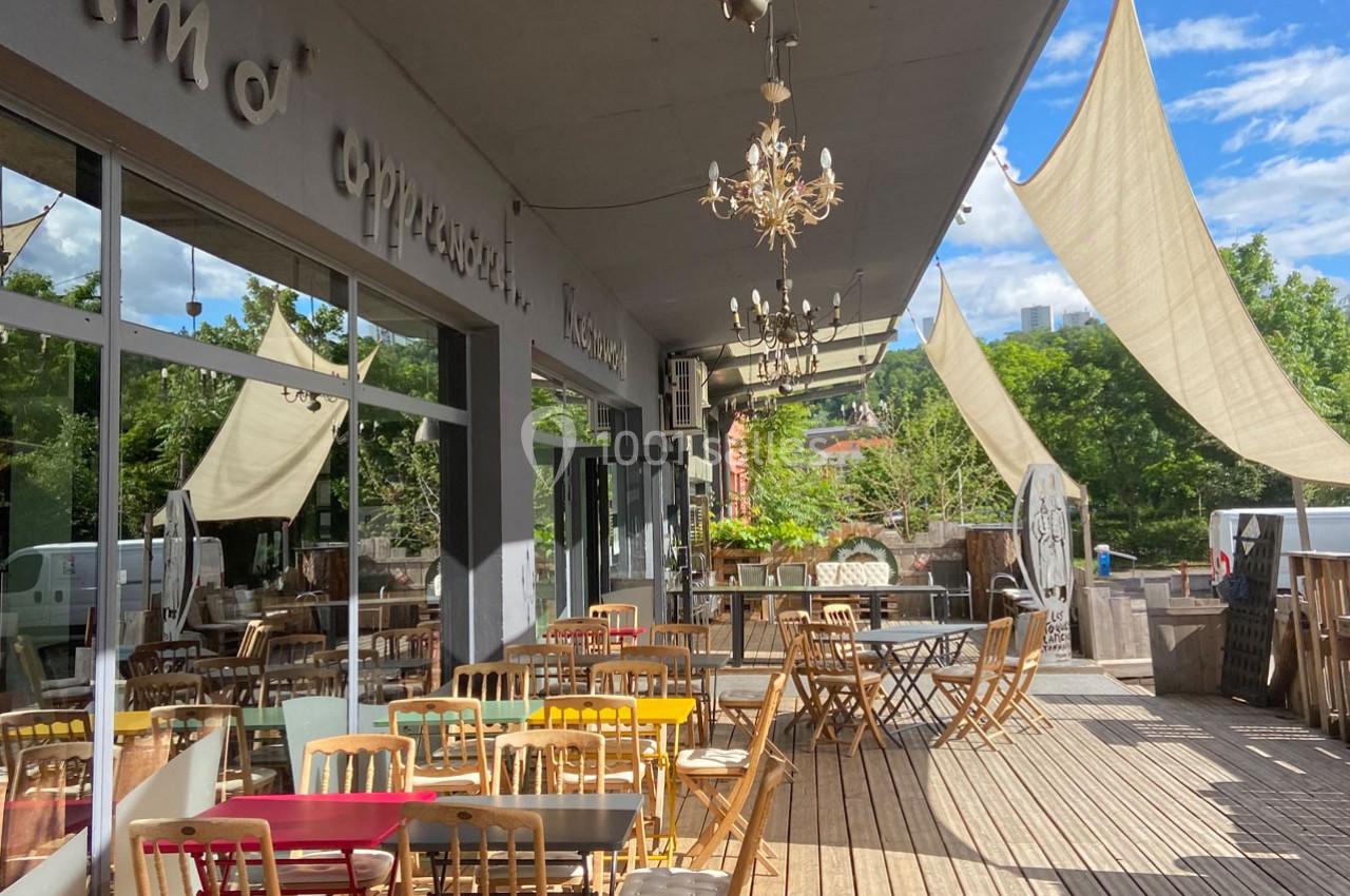Terrasse en bois d'un café avec des tables et chaises colorées, ombragée par des voiles tendues, entourée de verdure.