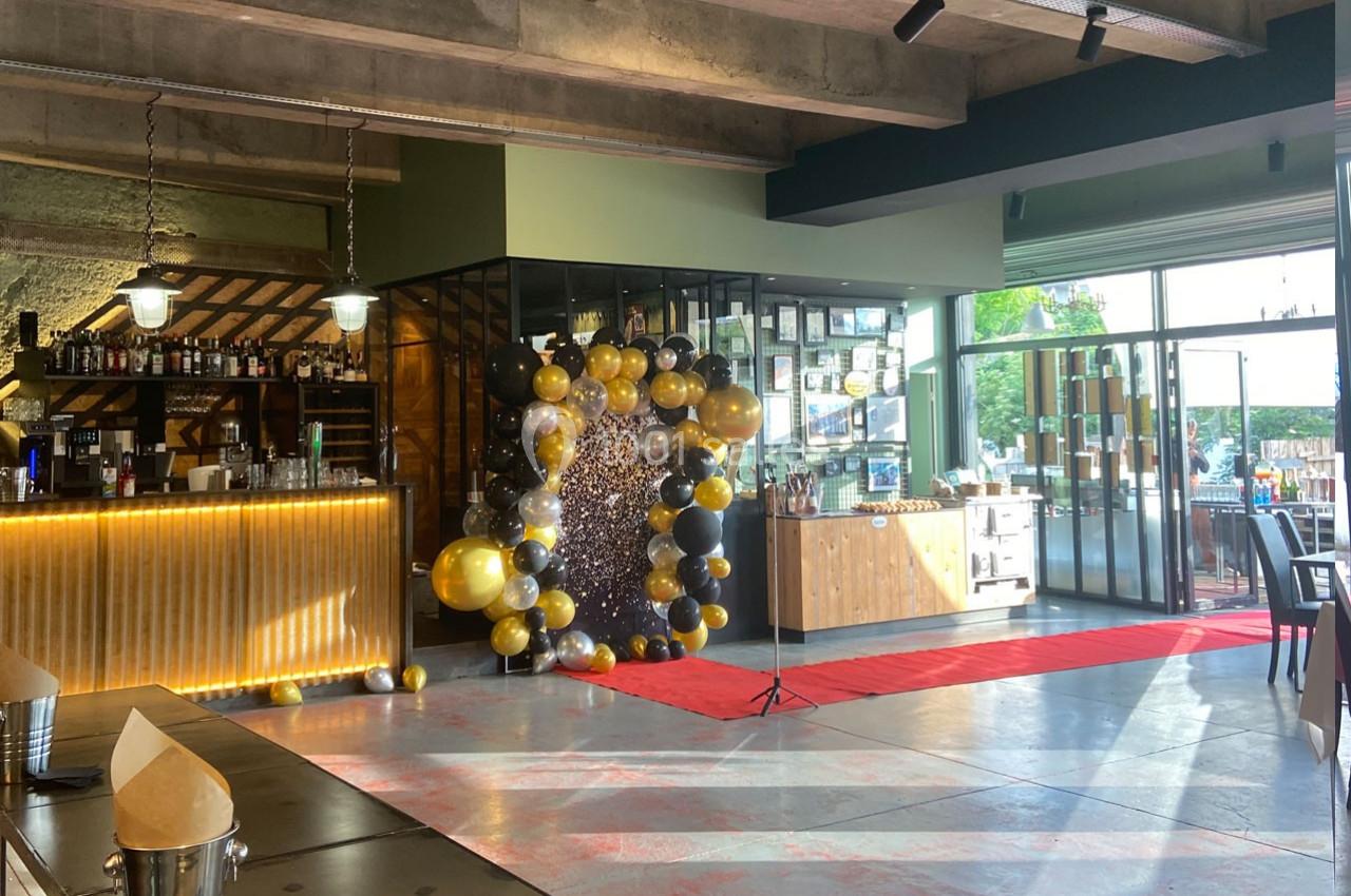 Salle de réception avec un bar éclairé, une arche de ballons dorés et noirs, et un tapis rouge près d'une baie vitrée.