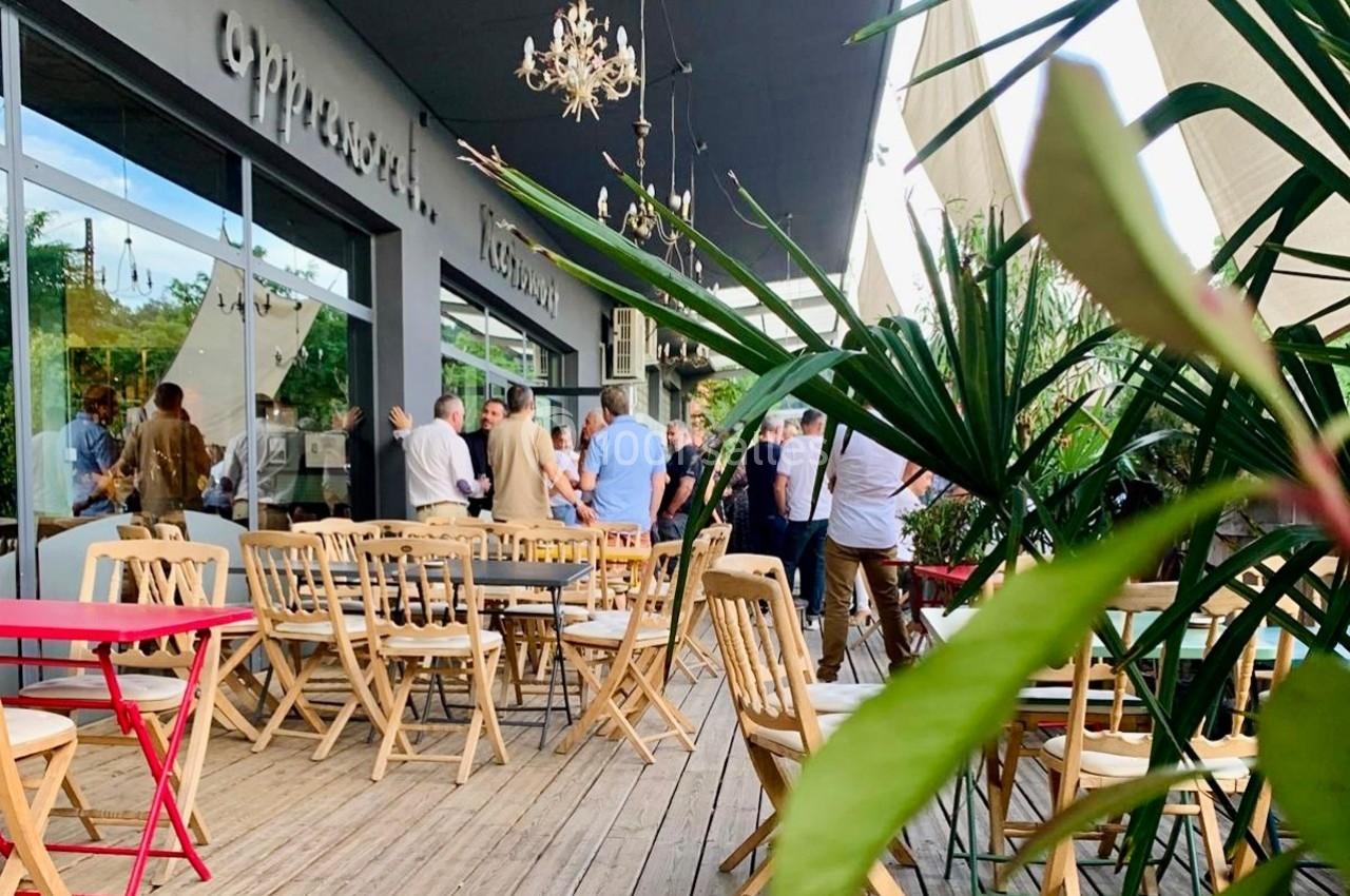 Terrasse en bois avec des tables et chaises, des plantes en premier plan, et un groupe de personnes discutant au fond.