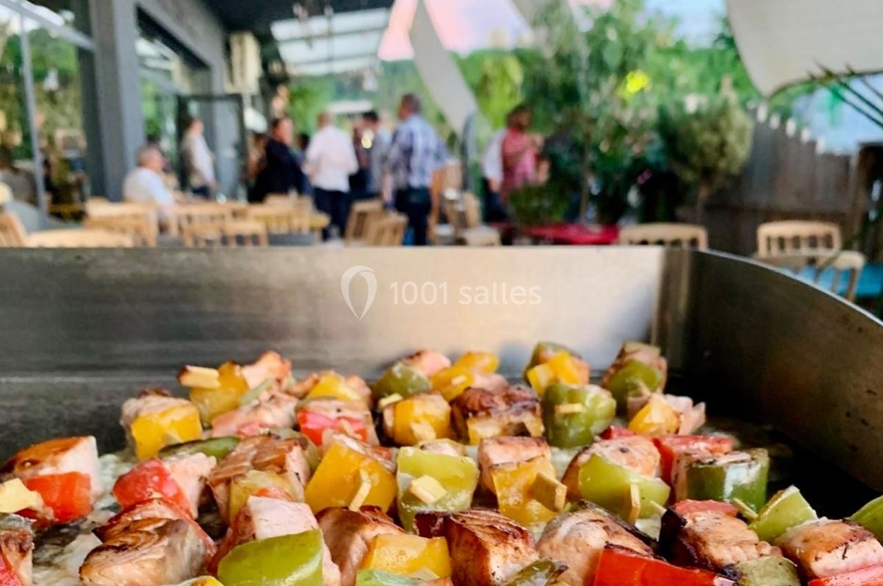 Brochettes de légumes et de viande grillées sur une plancha, avec une terrasse animée en arrière-plan.