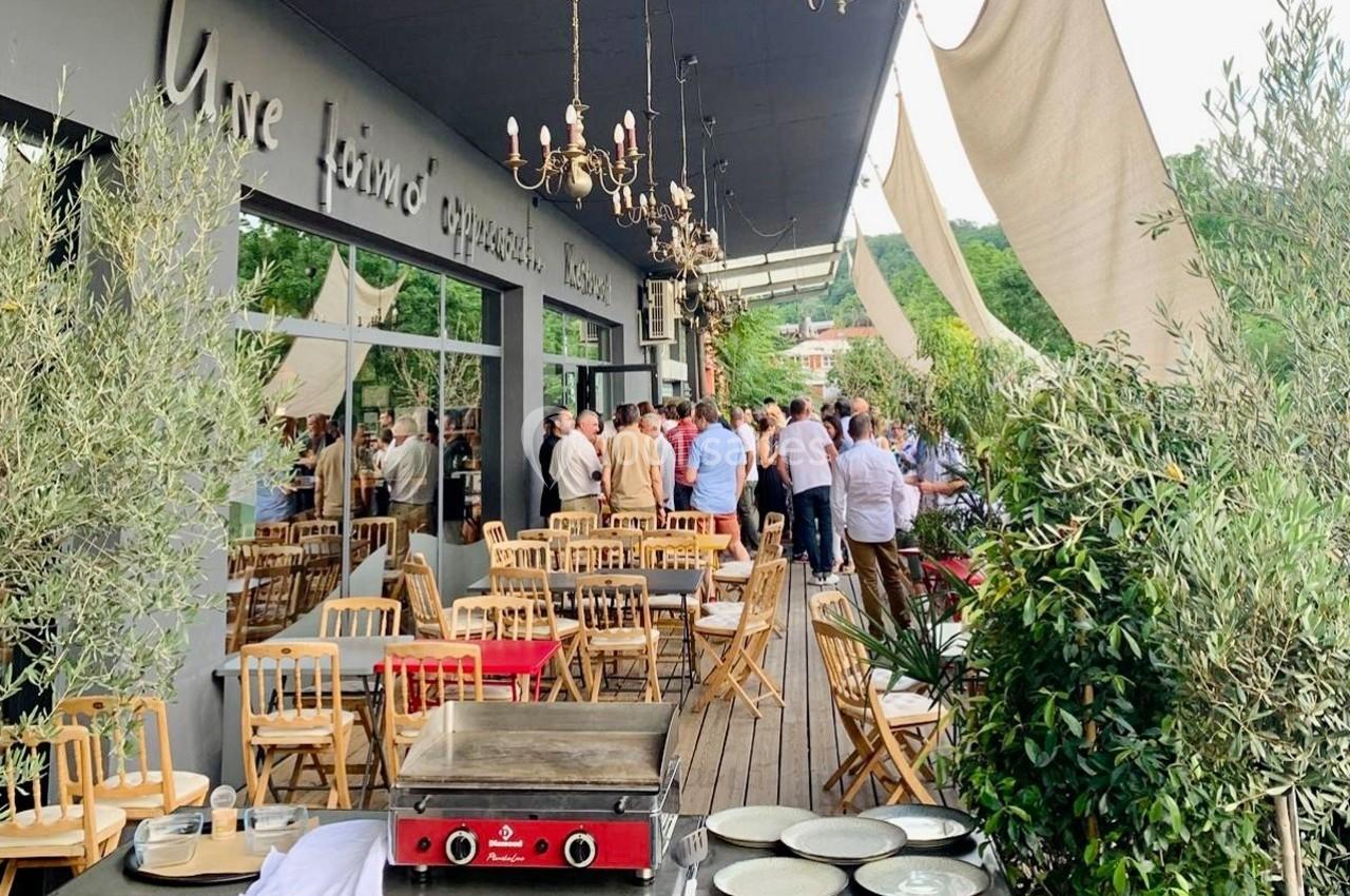 Terrasse d'un restaurant avec des tables en bois, des convives debout et des oliviers en décoration.