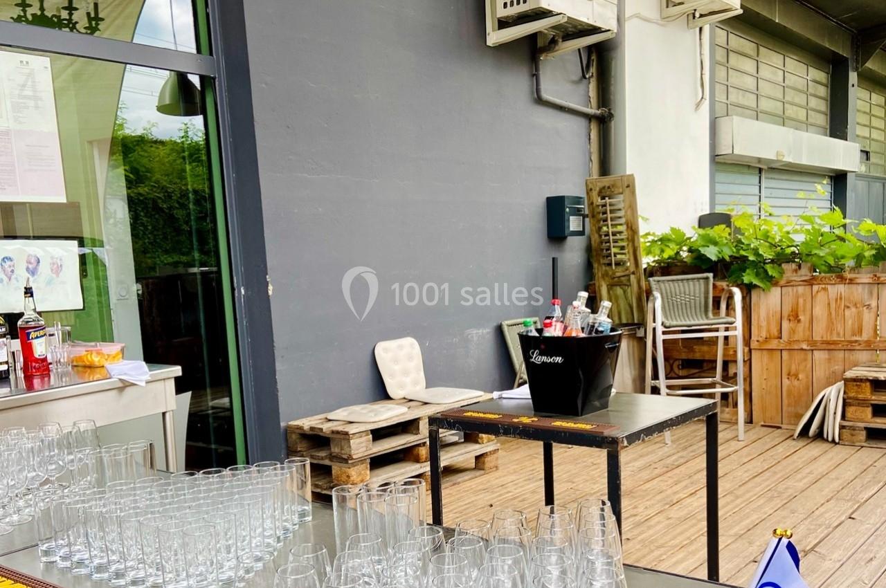 Terrasse aménagée avec des tables, des verres alignés, un seau à glace et des boissons, près d'un mur gris.