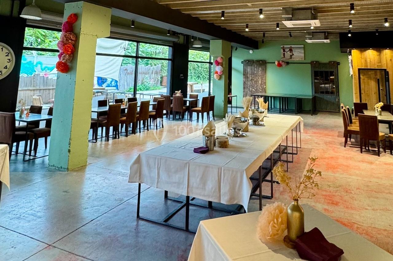 Salle de réception lumineuse avec tables décorées, chaises alignées et grandes baies vitrées donnant sur un espace extérieur.