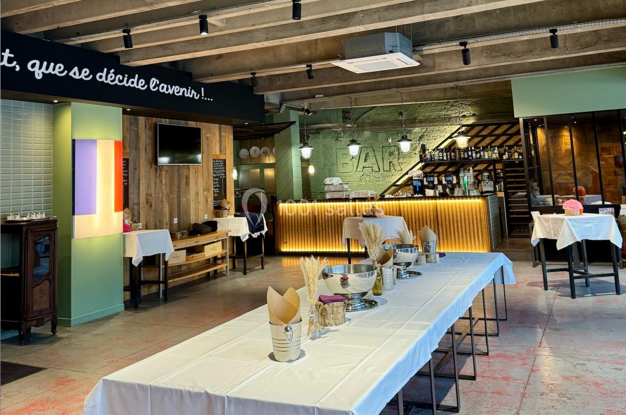 Salle de restaurant moderne avec tables dressées, bar éclairé en arrière-plan et ambiance industrielle.