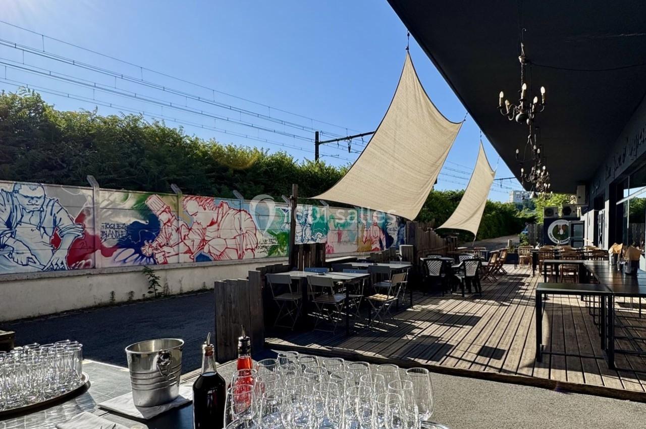 Terrasse extérieure avec tables, chaises, voiles d'ombrage et fresques colorées sur un mur longeant des voies ferrées.