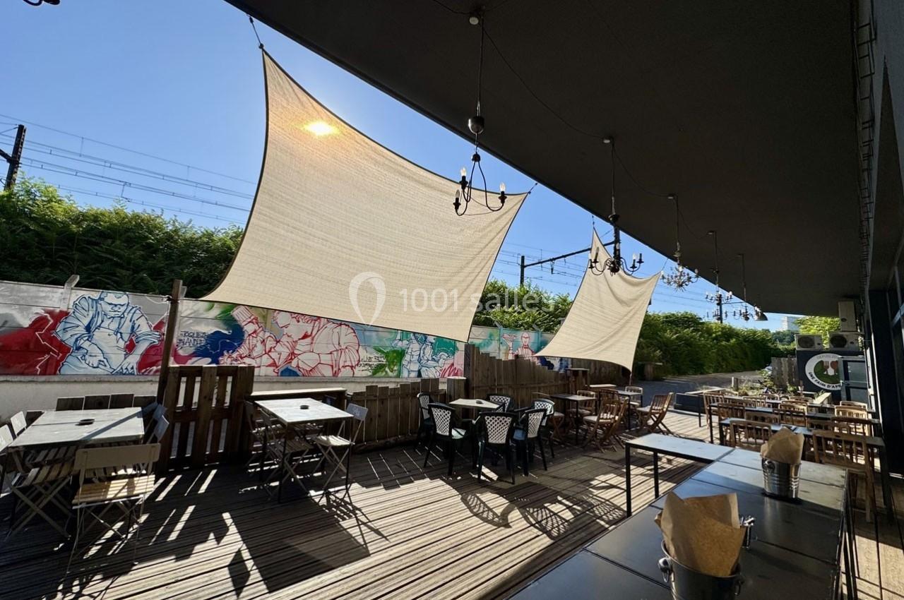 Terrasse en bois avec tables et chaises, ombragée par des voiles tendues, entourée de murs décorés de fresques colorées.