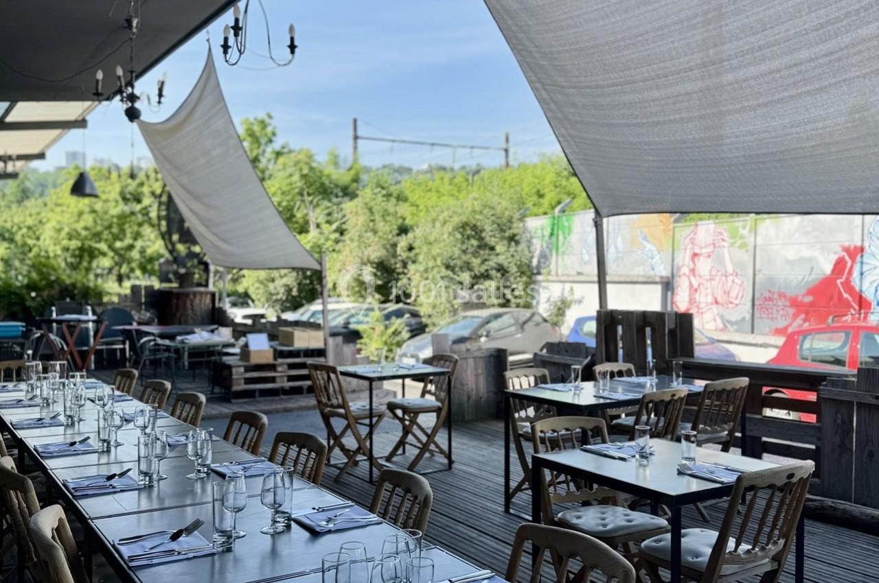 Terrasse ombragée avec de longues tables dressées, chaises en bois et vue sur un mur avec des graffitis colorés.