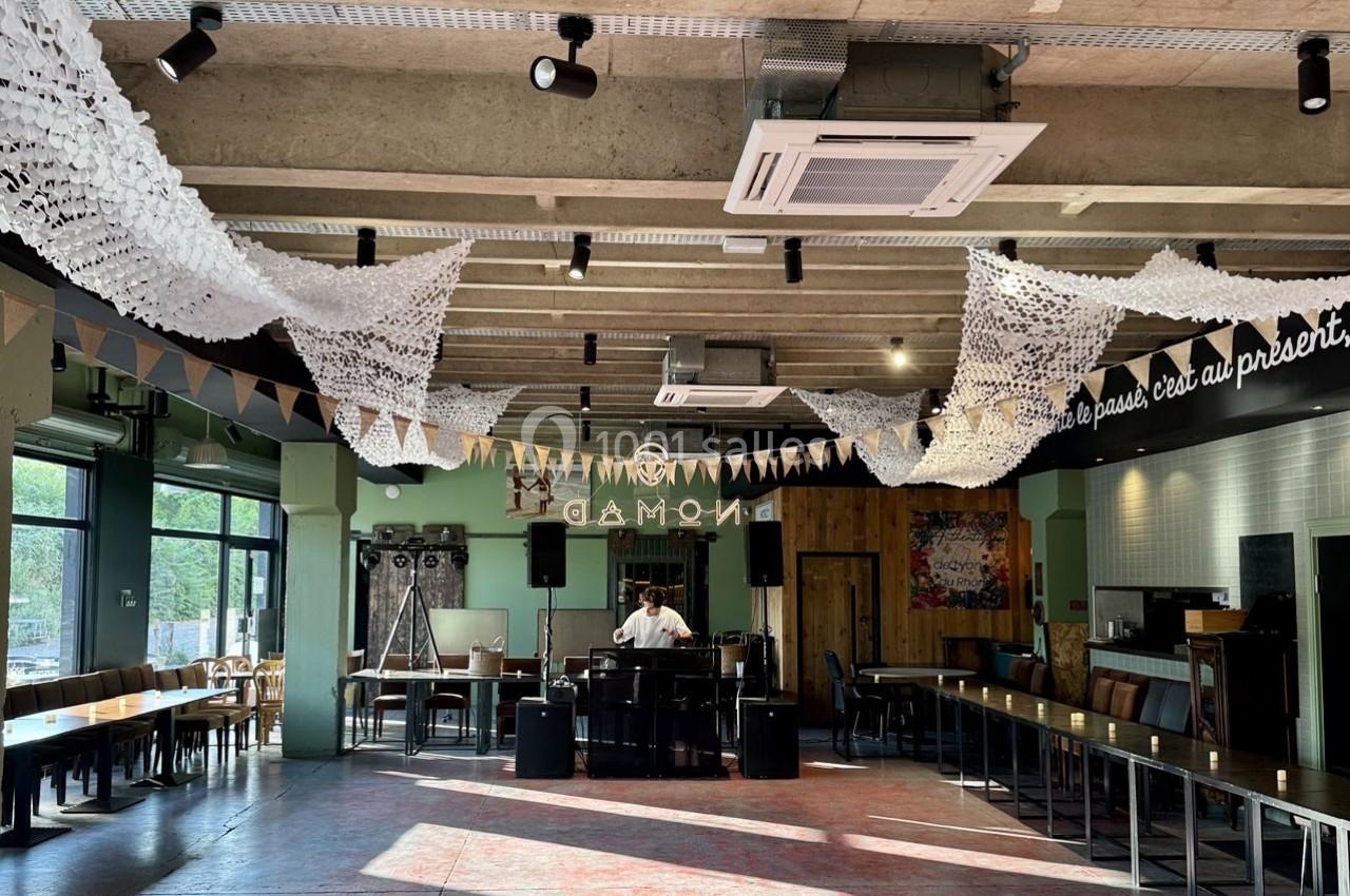 Salle lumineuse avec tables alignées, décorée de guirlandes et filets suspendus, avec un DJ au centre.