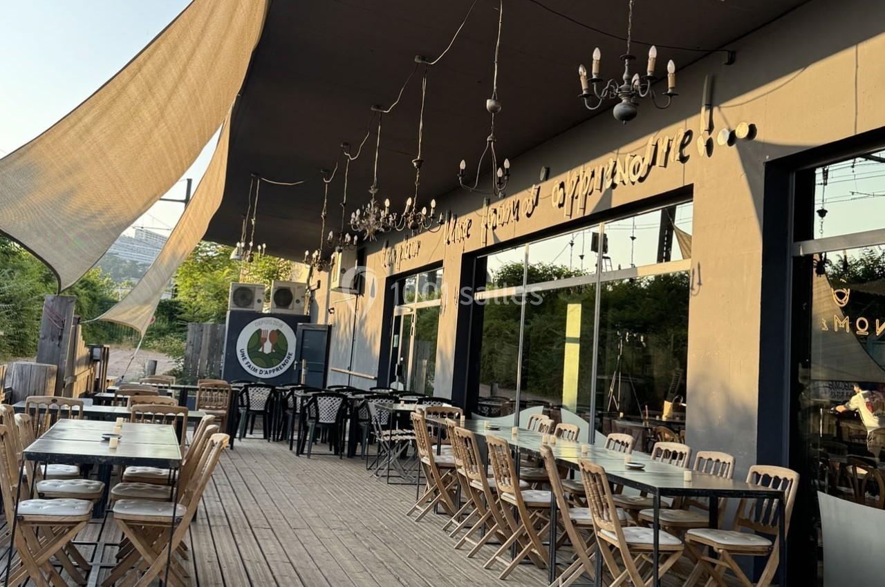 Terrasse en bois d'un restaurant avec des tables et chaises en extérieur, éclairée par des lustres suspendus.
