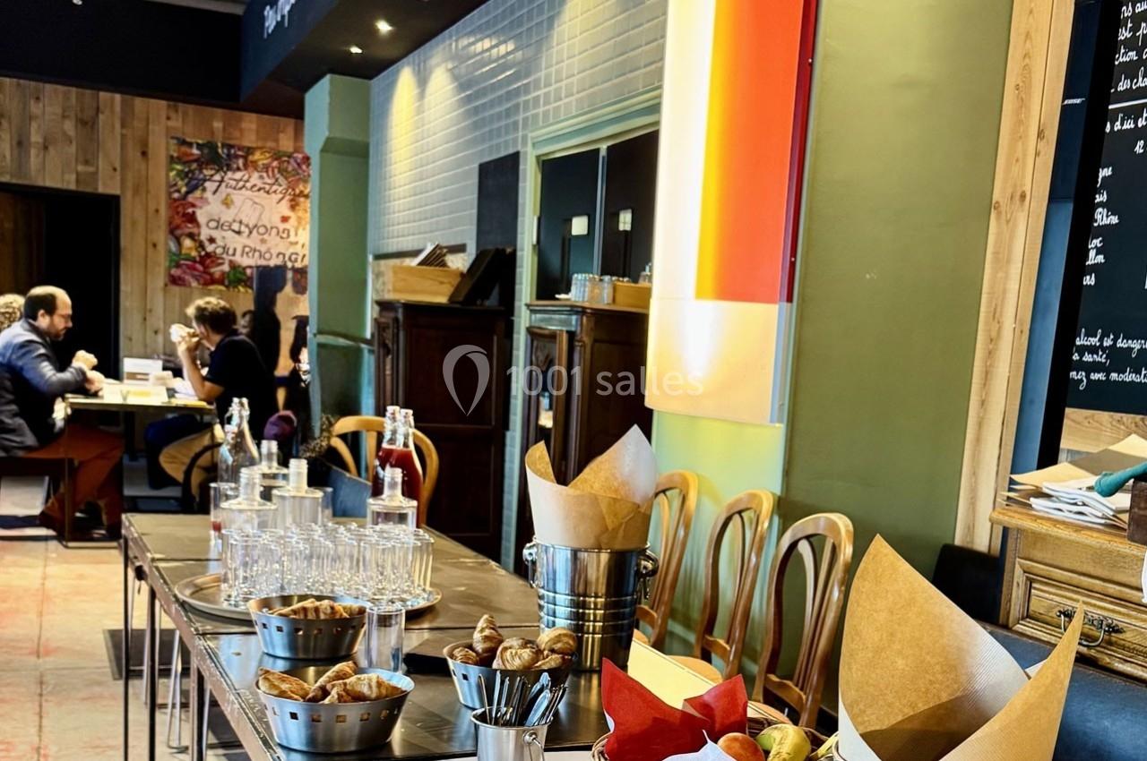 Table dressée avec viennoiseries, verres et carafes dans un café-restaurant avec des clients en arrière-plan.