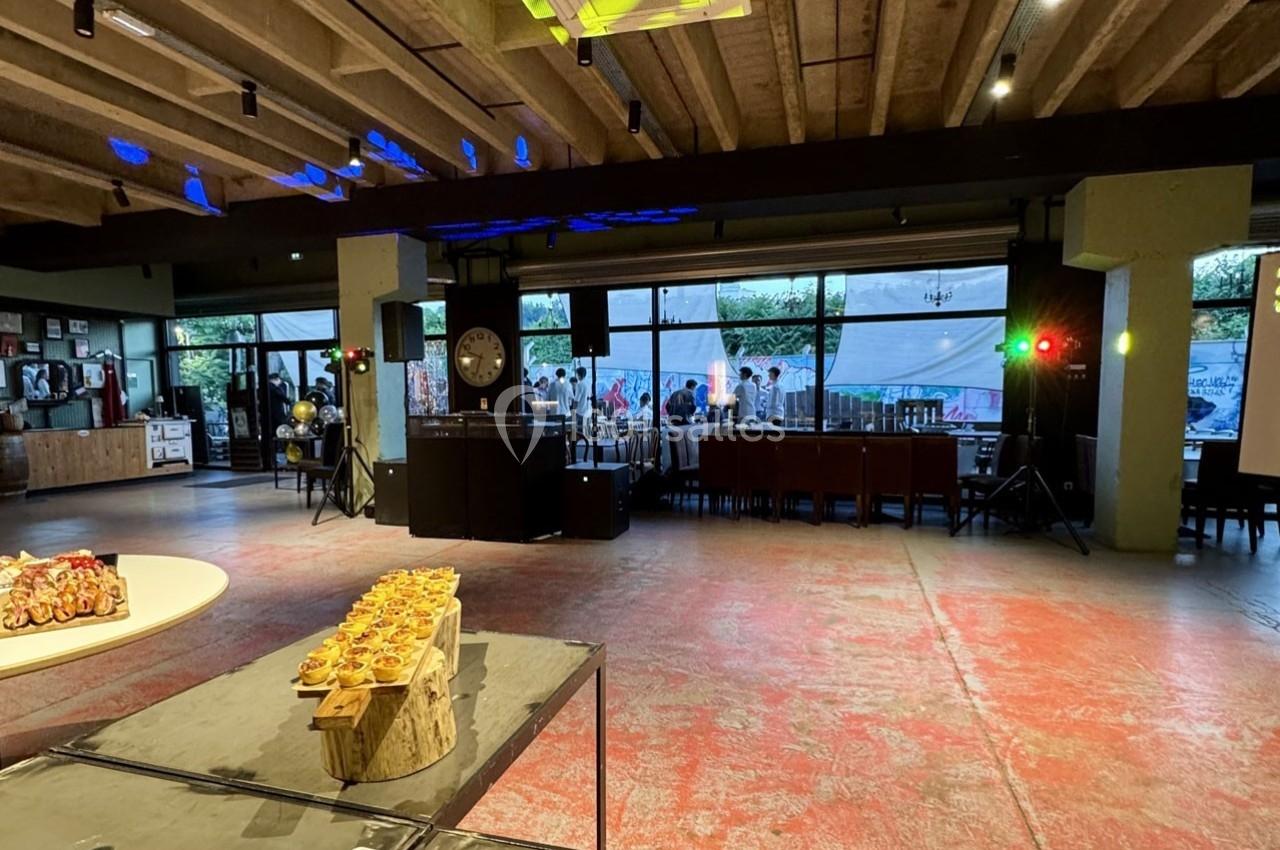 Salle spacieuse avec tables et buffet en premier plan, grandes baies vitrées donnant sur une terrasse animée en arrière-plan.