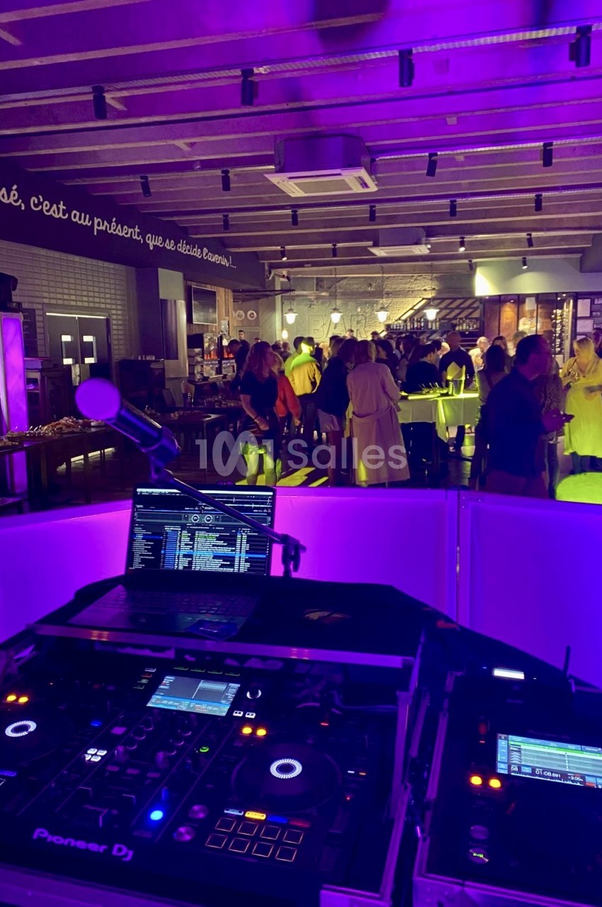 Salle de réception éclairée en violet avec console DJ au premier plan et convives attablés au bar en arrière-plan.