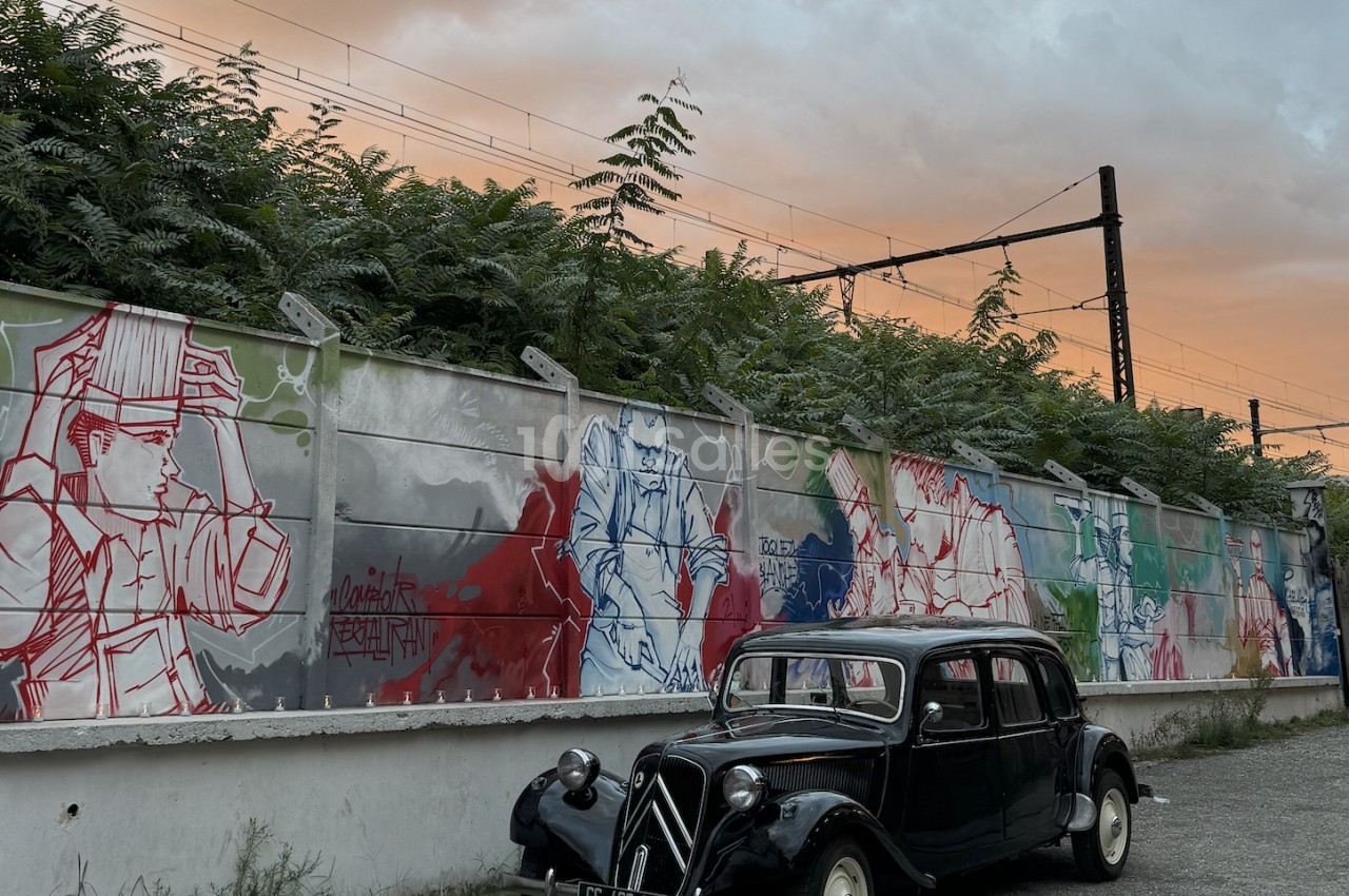 Voiture noire vintage stationnée devant mur orné de graffitis rouges et bleus, fond de ciel crépusculaire.
