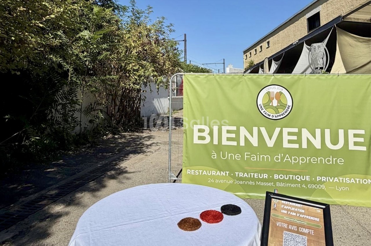 Espace extérieur avec table ronde dressée de blanc, bannière verte de bienvenue et bâtiment en arrière-plan sous ciel bleu.