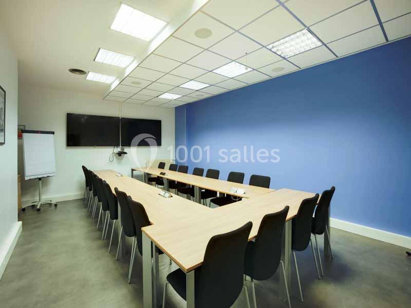 Salle de réunion lumineuse avec tables disposées en U, chaises noires, mur bleu et écran mural.
