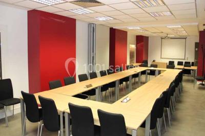 Salle de réunion avec table en bois, chaises noires, mur rouge décoré et écran mural éteint.