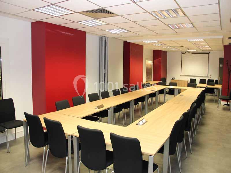 Salle de réunion avec tables disposées en U, chaises noires, murs rouges et un projecteur au fond.