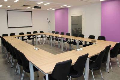 Salle de réunion avec table en bois, chaises noires, mur rouge décoré et écran mural éteint.