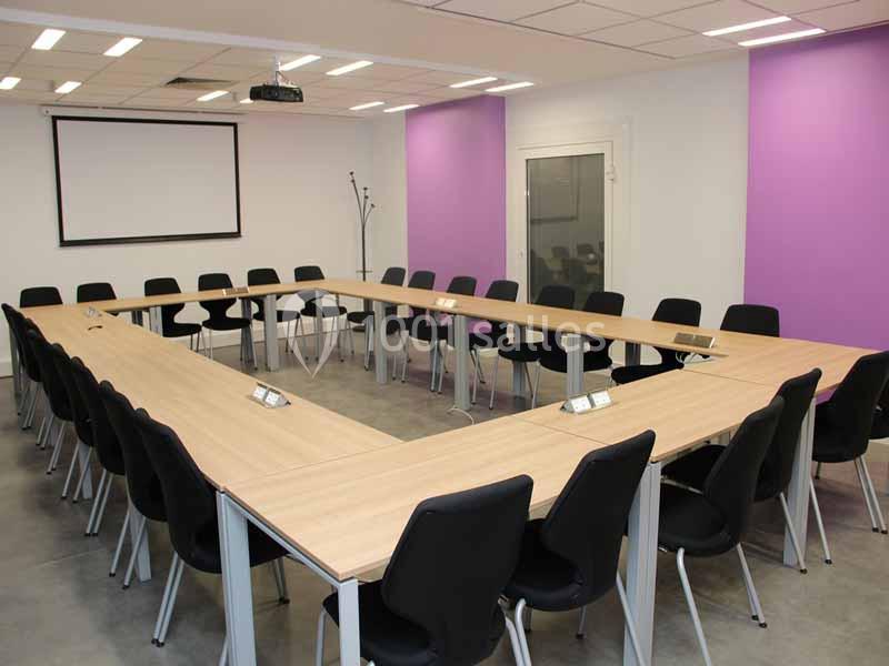 Salle de réunion avec tables disposées en U, chaises noires, écran de projection et murs partiellement violets.