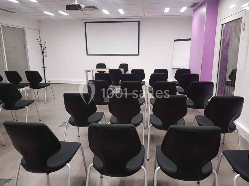 Salle de réunion avec des rangées de chaises noires, un écran de projection, un tableau blanc et des murs violets.