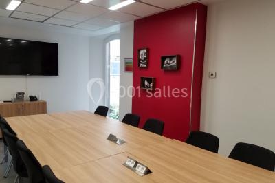 Salle de réunion avec table en bois, chaises noires, mur rouge décoré et écran mural éteint.