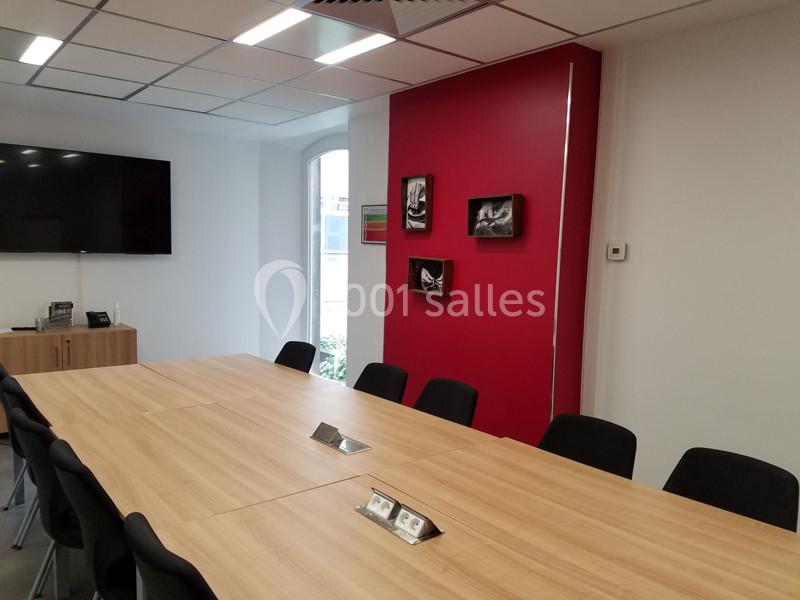 Salle de réunion moderne avec grande table en bois, chaises noires, écran mural et décorations sur un mur rouge.