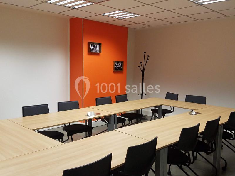 Salle de réunion avec tables en U, chaises noires, mur orange décoré de cadres et porte-manteau dans un coin.