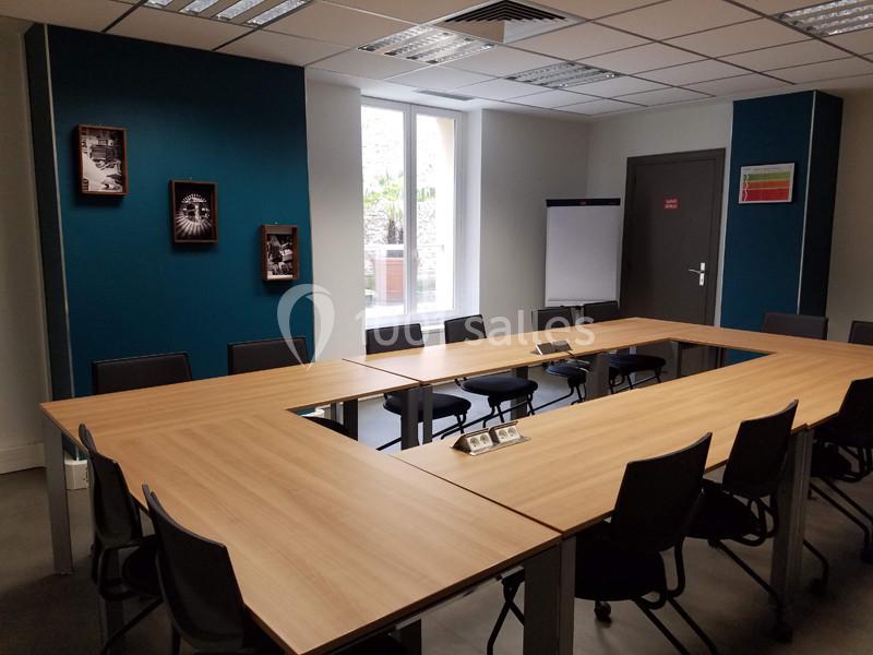 Salle de réunion lumineuse avec table en U, chaises noires, tableau blanc et décorations murales.