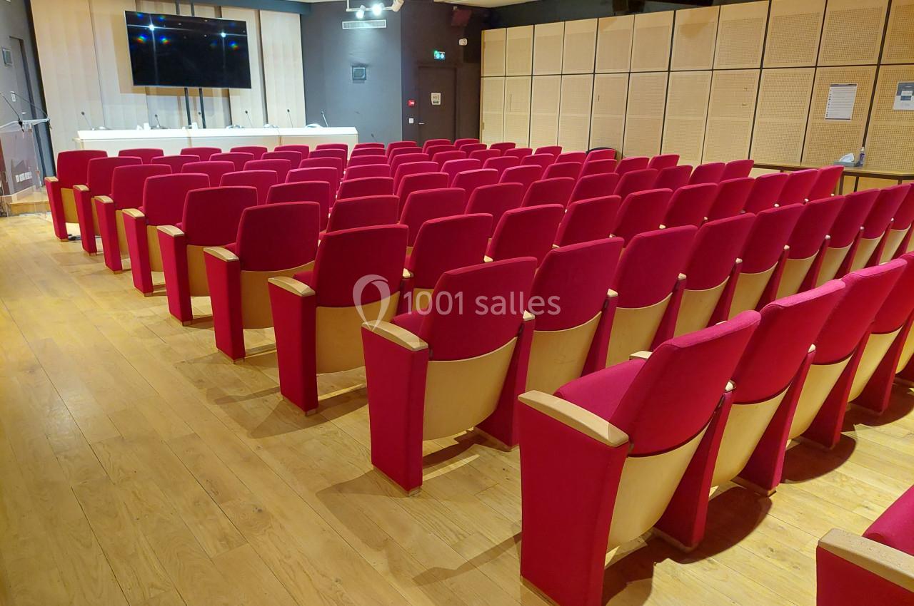 Salle de conférence avec rangées de fauteuils rouges, parquet en bois et écran mural à l'avant.