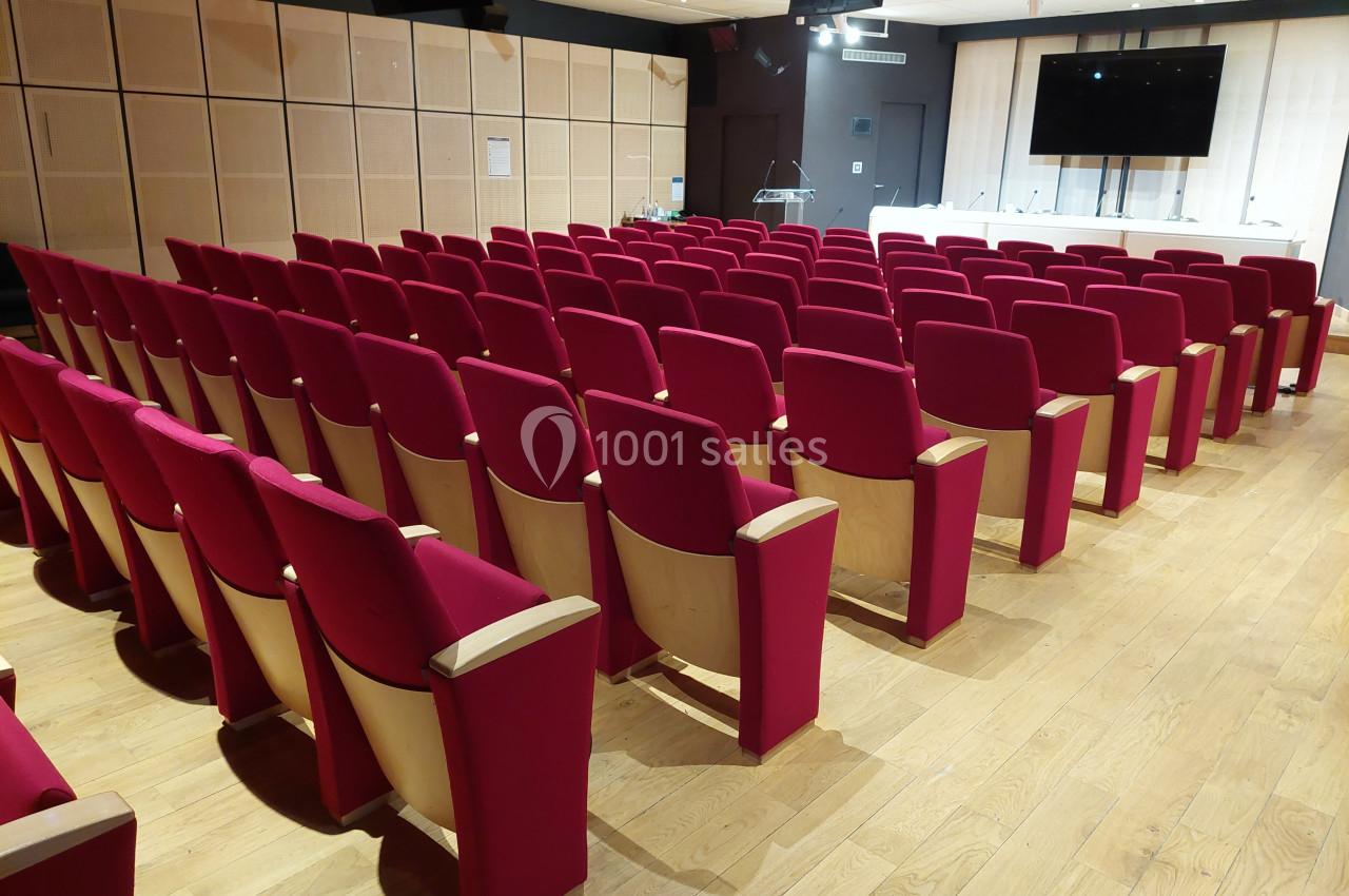 Salle de conférence avec rangées de sièges rouges en bois, écran mural et estrade à l'avant.