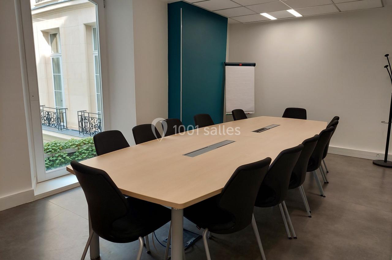 Salle de réunion lumineuse avec une grande table rectangulaire entourée de chaises noires et un paperboard au fond.