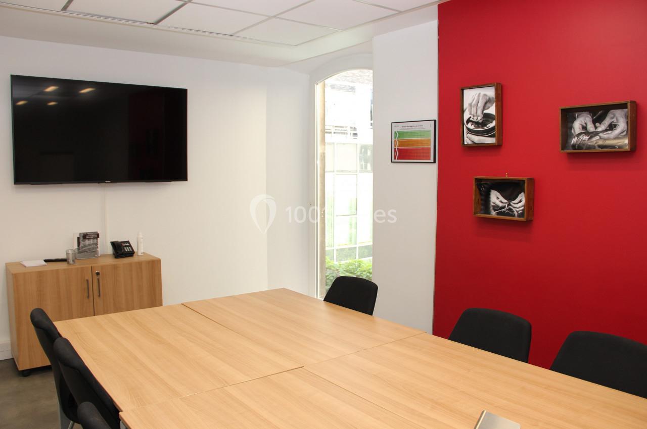 Salle de réunion avec table en bois, chaises noires, mur rouge décoré et écran mural éteint.