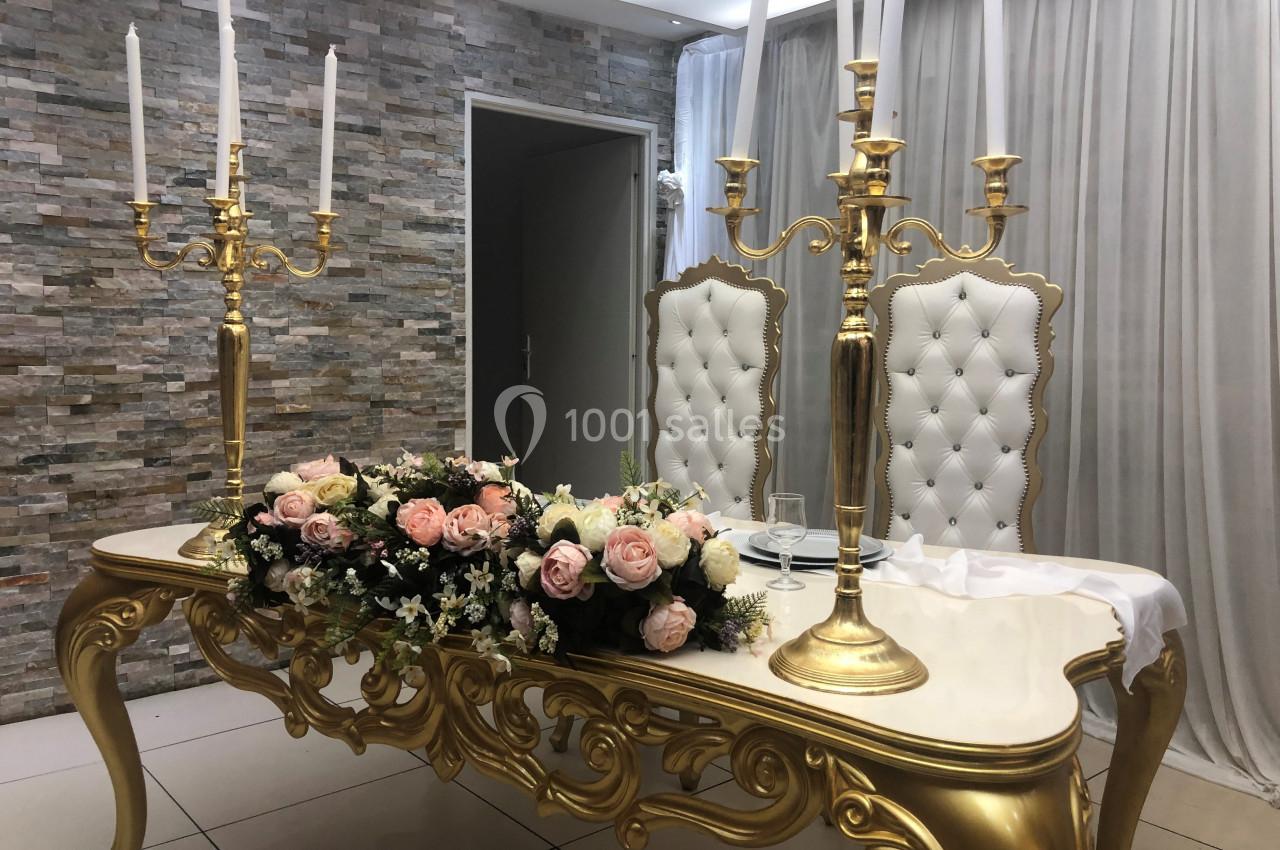 Table ornée de dorures avec chandeliers, fleurs roses et blanches, deux chaises capitonnées blanches en arrière-plan.