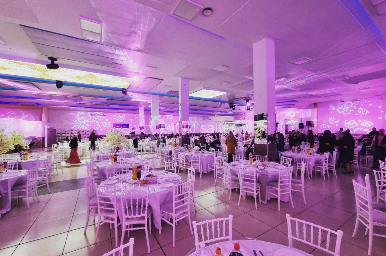 Salle de réception décorée avec des tables rondes, nappes blanches, chaises blanches et éclairage rose.